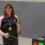 A woman kneels indoors holding a trophy and fencing sword, wearing a medal. A digital scoreboard behind her shows a score of 15:14 with a time of 3:00.