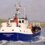 Small passenger and vehicle ferry named Good Shepherd IV, Lerwick, sails across calm water near a grassy shoreline.
