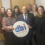 Seven people stand indoors, smiling at the camera. One person in front holds a sign that reads “dbi Distress Brief Intervention: Connected Compassionate Support.”.