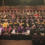 A large group of people, many in academic gowns, are seated in an auditorium for what appears to be a graduation or formal ceremony.