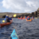 People kayaking in a line on open water near rocky cliffs, with a cloudy sky and hilly landscape in the background.