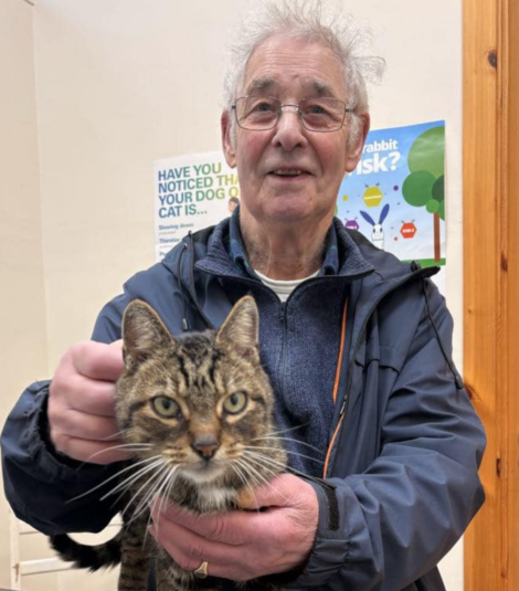 An older man in a jacket holds a tabby cat indoors. Posters about pet care are on the wall behind them.