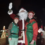 Santa and two elves wave from a festively lit vehicle at night.
