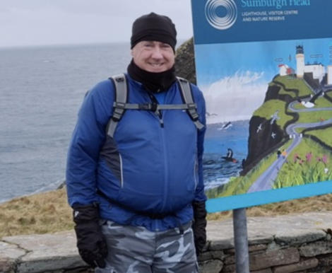 Person standing outdoors near a map sign, dressed in a blue jacket, black gloves and hat, with a backpack. Coastal scenery and ocean visible in the background.