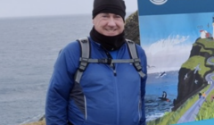 A person in a blue jacket and black hat stands outdoors near a nature reserve sign, with the ocean in the background.