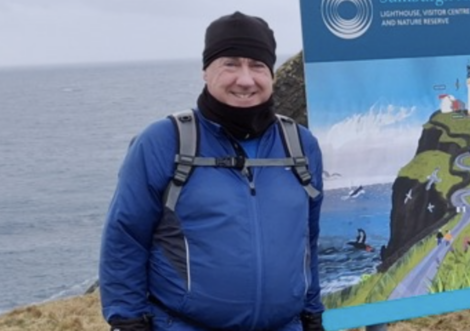 A person in a blue jacket and black hat stands outdoors near a nature reserve sign, with the ocean in the background.