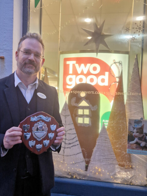 Man in a suit holding a shield-shaped plaque in front of a decorated window with a "Two good" sign.