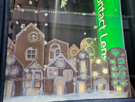 Gingerbread-style houses with white icing details are displayed in a shop window. Snowflakes and a green sign with "contact lenses" are visible.