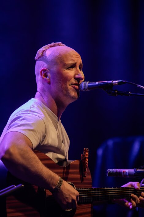 A man with a bald head and a patch plays an acoustic guitar and sings into a microphone on stage under blue and purple lights.