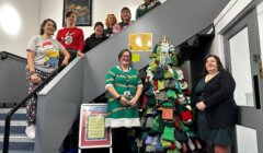A group of people in festive clothing pose beside a Christmas tree made of knit hats on a staircase indoors.