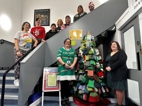 A group of people in festive clothing pose beside a Christmas tree made of knit hats on a staircase indoors.