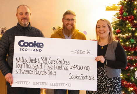 Three people hold a large check for £4,520.00 from Cooke Scotland to Walls, Unst & Yell Care Centres. A Christmas tree is in the background.