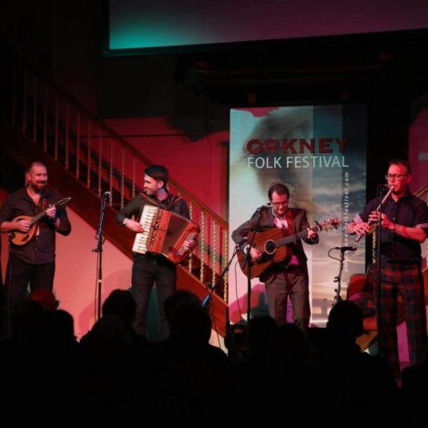 A band performs on stage at the Orkney Folk Festival, featuring musicians playing a mandolin, accordion, guitar, and woodwind instrument.