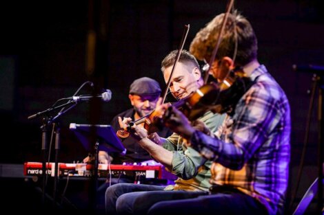 Three musicians performing on stage with violins and a keyboard.