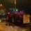 Several tractors decorated with Christmas lights drive down a wet street at night. A sign on the front tractor reads "Christmas Tractor Run.