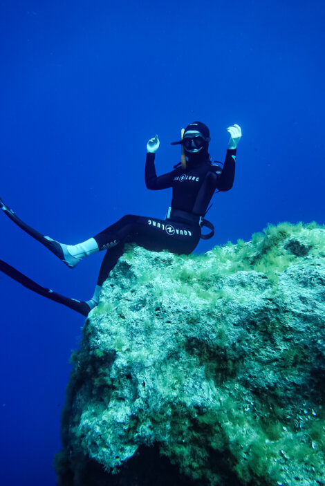 A diver in a wetsuit and fins sits on a large underwater rock, surrounded by blue water and green algae.