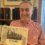 Man holding a "75 Years of the SFA" book, standing in front of a bookshelf and window.
