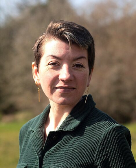Person with short hair and earrings stands outdoors, wearing a dark green jacket.