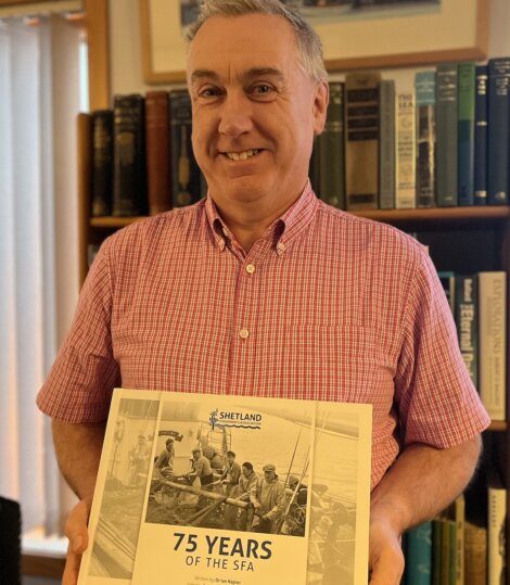 A man in a red checkered shirt smiles while holding a book titled "75 Years of the SFA" with a historical photograph on the cover.