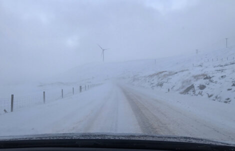 Snow-covered road with a wind turbine in the background, surrounded by a snowy landscape and overcast sky.