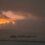 A rural landscape shows a snow-covered field with a farm in the distance and a dramatic orange sunset in a cloudy sky.