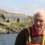 An older man stands by a lake with rocky terrain and stone houses in the background under a cloudy sky.