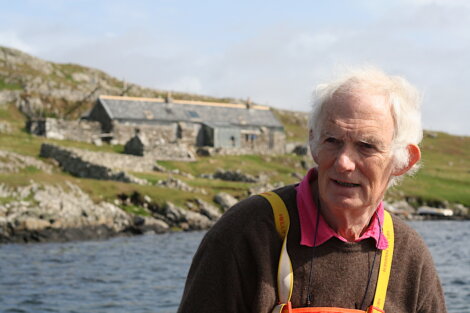 An older man stands by a lake with rocky terrain and stone houses in the background under a cloudy sky.