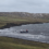 A small boat with a red crane is anchored close to the rocky shore of a grassy, hilly coastline under a cloudy sky.