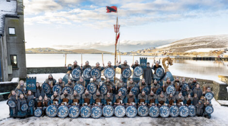 A large group of people in Viking attire hold blue shields, posing outdoors on a snowy day. A flag and a snowy landscape are visible in the background.