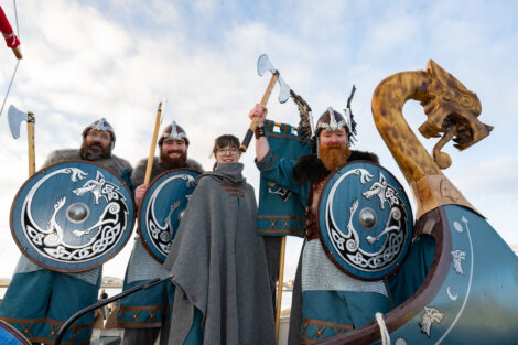 A person stands with four individuals dressed as Viking warriors holding shields and axes, next to a boat with a dragon head prow.