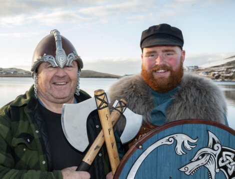 Two men dressed in Viking-inspired outfits, one wearing a helmet, the other a fur cloak, holding battle axes and a shield, standing by a lake with hills in the background.