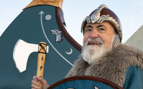 Elderly man dressed as a Viking, holding an axe, standing in front of a decorated shield or ship.