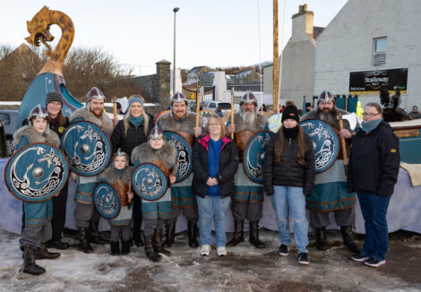 A group of people in Viking costumes with shields pose alongside others in winter clothing. A large decorative vehicle is in the background.