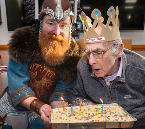 An elderly man wearing a "Birthday Boy!" crown blows out candles on a cake. A person in Viking attire poses beside him, holding the cake.