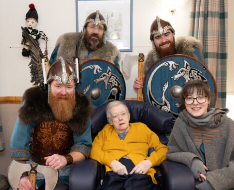 A seated elderly woman is surrounded by three people in Viking costumes with helmets and shields, and a woman wearing glasses. They are indoors, smiling for a group photo.