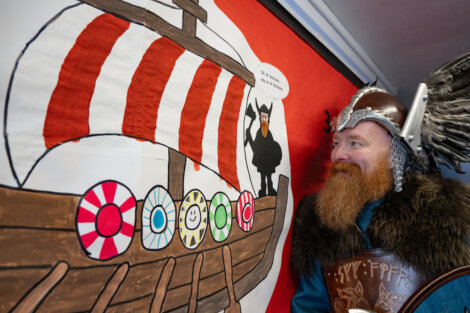 Man dressed as a Viking stands next to a wall display featuring a Viking ship and a cartoon figure.