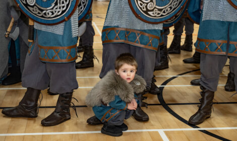 A young child in medieval-style clothing crouches on a wooden floor, surrounded by adults in similar attire, holding shields.