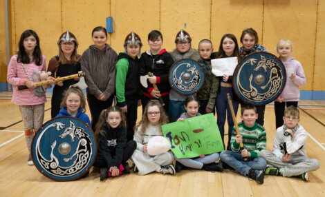 A group of children poses in a gym wearing Viking helmets, holding shields and signs.