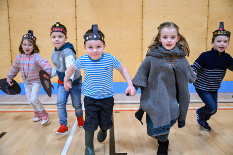 Five children, wearing medieval-style costumes and headbands, run indoors on a wooden floor.
