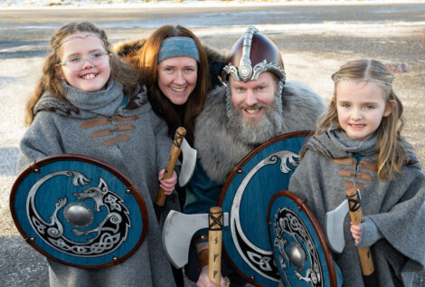 A family wearing Viking costumes, including helmets, shields, and axes, poses together outdoors, smiling at the camera.