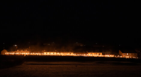 Row of houses with lit torches outside, creating a line of light against the night sky.