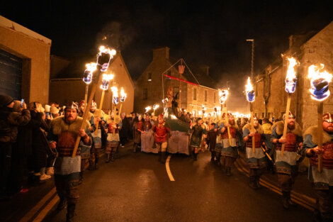 Participants in Viking costumes carry torches during a nighttime parade on a street lined with spectators.
