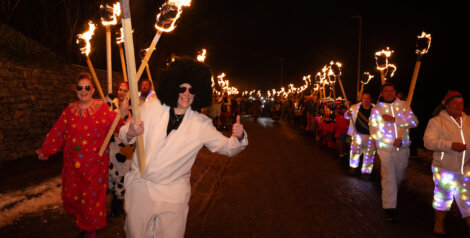A group of people in colorful costumes march at night holding flaming torches, with one person giving a thumbs-up in the foreground.