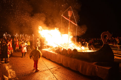 A Viking longship model is set ablaze during a nighttime outdoor event, surrounded by people in Viking costumes holding torches.