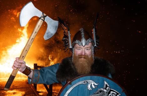 Person in Viking costume holding an axe and shield, standing in front of a large fire at night.