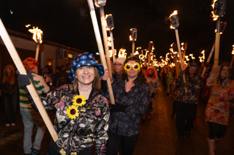 A group of people wearing floral patterns and holding torches walk in a nighttime parade. Two women in focus have sunflowers pinned to their jackets.