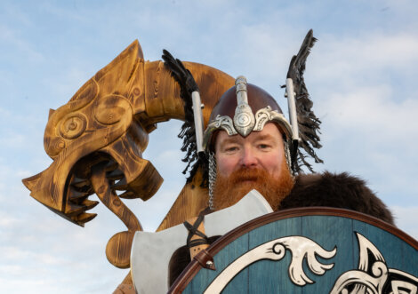 Person in Viking warrior attire with helmet and axe, positioned in front of a large wooden dragon sculpture, under a blue sky.