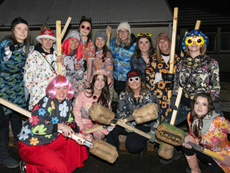 Group of people in colorful floral clothing and wigs, holding large wooden mallets, pose together in front of a building at night.