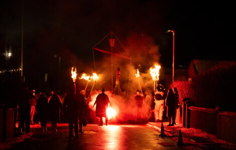 People walk at night holding torches, surrounding a small boat structure. The scene is illuminated with red and orange light. A smoky atmosphere adds to the dramatic effect.