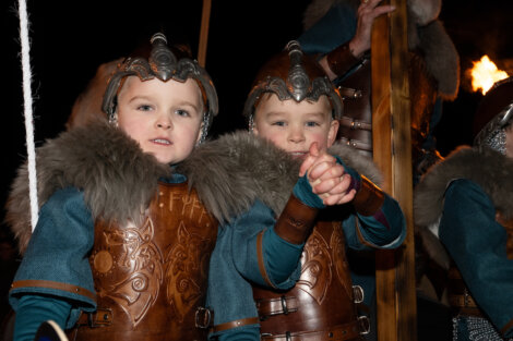 Two children in Viking costumes with helmets and fur-trimmed capes hold hands.